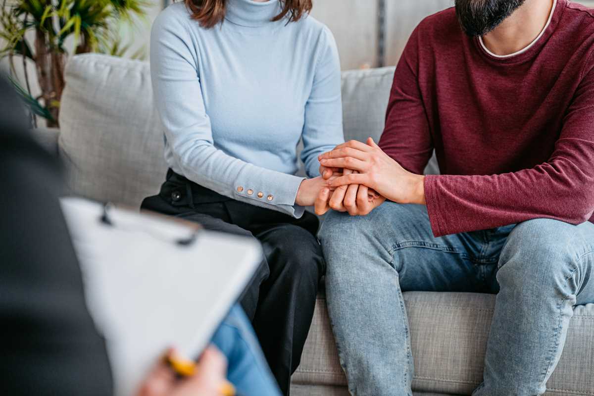 Ein Paar (deren Gesichter nicht erkennbar sind) sitzen nebeneinander und halten sich die Hand. Gegenüber sitzt eine verschwommene Person mit Klemmbrett.